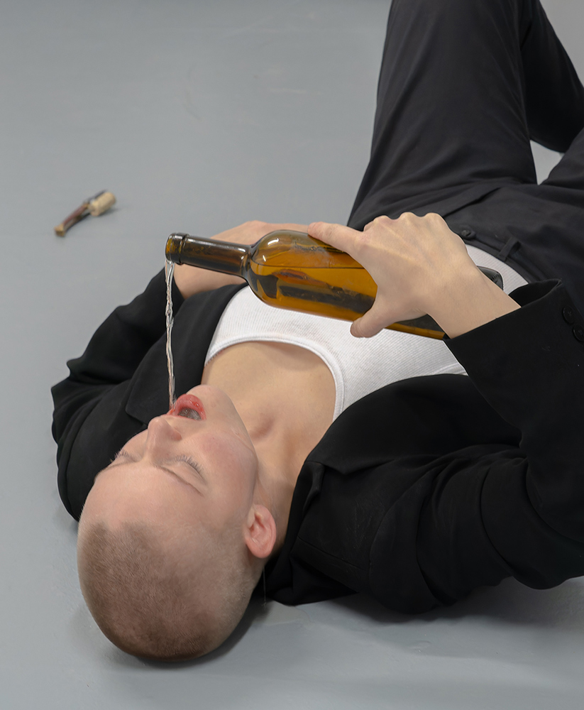 The artist is laying on the ground. They are pouring clear liquid into their mouth from what seems to be an old wine bottle.